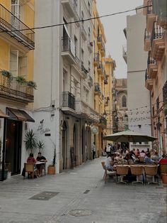 people are sitting at tables in the middle of an alleyway with buildings and balconies