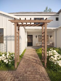 a brick walkway leading to a white building with flowers in the foreground and a wooden pergolated arbor