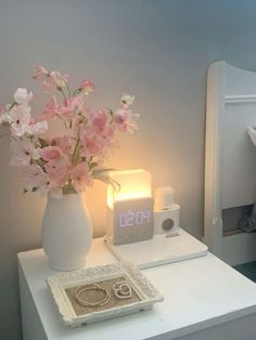 a white vase with pink flowers sitting on top of a table next to a clock