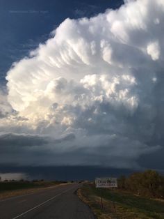 a large cloud looms over a road in the middle of an open field with a sign on it