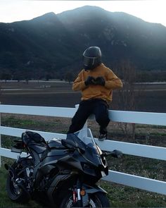 a man sitting on top of a motorcycle next to a white fence with mountains in the background