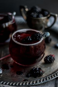 a glass filled with liquid and blackberries on top of a metal tray next to two silver cups