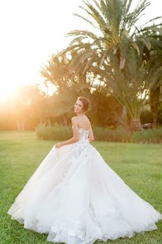 a woman in a wedding dress is standing on the grass with palm trees behind her