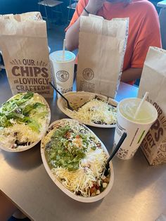 two burritos and drinks sitting on a table with paper bags in front of them