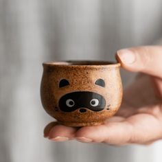 a hand holding a small ceramic cup with a racoon face painted on it