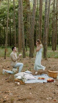 a man kneeling down next to a woman on a blanket in the middle of a forest