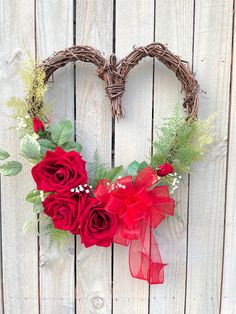 a heart shaped wreath with red roses and greenery hanging on a wooden fence behind it