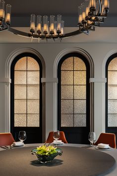 a round table with orange chairs in front of three arched windows and chandelier