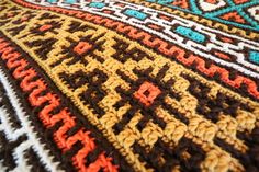 a crocheted blanket is laying on top of a tablecloth with an orange, brown, and blue design