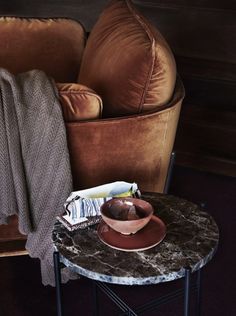 a table with a bowl on it next to a brown chair and throw blanket in the background