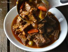 a white bowl filled with meat and vegetables on top of a wooden table next to a spoon