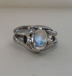 The Crystal Ball Ring is a very unique design, crafted by Heirloom Pavé in 18K white gold, featuring a Cabochon Moonstone. And two perfectly blue round brilliant Sapphires. The Moonstone is a oval shape Cabochon with a beautiful iridescent that so mystical and other worldly looking. The two, bezel-set Sapphires are a total carat weight of .35ct are a beautiful accent to the center. In the high polished white gold it is a true statement piece. Size 7 (Sizable upon request) Every Antique piece of Unique White Multi-stone Moonstone Ring, Unique Opal Ring With Moonstone, Fusion Style Moonstone Ring Gift, Unique Silver Moonstone Crystal Ring, Mystical Cabochon Ring For Anniversary, Handmade Celestial Silver Opal Ring, Mystical Anniversary Ring With Cabochon, Unique Moonstone Wedding Ring, Mystical Handmade Moonstone Wedding Ring