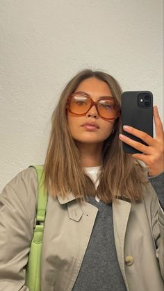 a woman wearing glasses taking a selfie with her cell phone while standing in front of a white wall