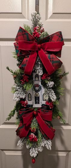 a wreath hanging on the front door decorated with red and green bows, pine cones, evergreens and a white house