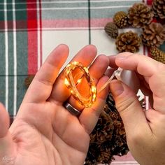 a person holding a ring with lights in their hands on a plaid table cloth next to pine cones