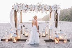 a woman standing on top of a sandy beach next to a wooden structure covered in white flowers