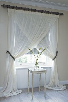 a vase with flowers sitting on a table in front of a window covered by sheer curtains