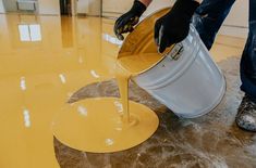 a man is pouring yellow paint on the floor with a white bucket and black gloves