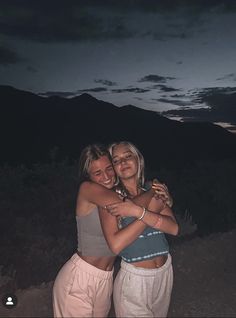 two young women hugging each other in front of a mountain at night with the sun setting behind them