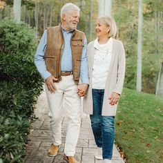 an older man and woman holding hands walking down a path in the park together on a fall day