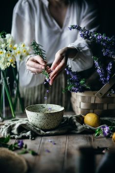 a woman arranging flowers in a bowl on a wooden table next to some lemons