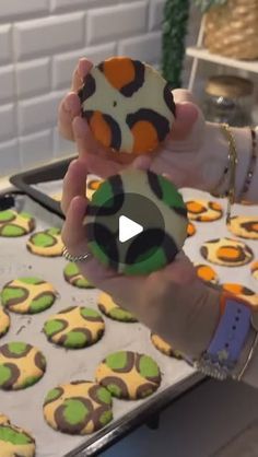 a person holding up some cookies in front of a pan full of decorated cupcakes