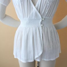 A stunning 1930s vintage blouse. Crisp, white cotton gauze with floral appliqué embroidery. Lap over short sleeves with plunging v-neck, elastic smocked waistband with double button and wrap front bust. All trim is lined with petite lace. Measurements of garment, allow space for fit.Taken flat, doubled for circumferenceShown on a size 4 dress formBust: 40”Waist: 22-28”Hips: 40”Shoulder: 16”Length: 24”Sleeve: 5.5”Tag: noneFabric: gauzy cottonCondition: Very good vintage. Buttons have been replace Vintage Summer Tops With Smocked Bodice, Vintage Smocked Bodice Top For Summer, Feminine Smocked V-neck Blouse, Fitted V-neck Coquette Tops, Fitted Smock V-neck Blouse, Fitted V-neck Smock Blouse, Vintage White Summer Blouse For Daywear, Vintage White Tops For Summer Daywear, V-neck Blouse With Smocked Bodice For Daywear