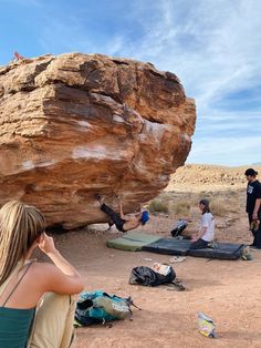 some people are sitting in the sand near a big rock