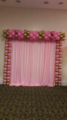 a pink and gold balloon arch in the middle of a carpeted room with curtains