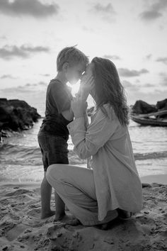 a woman kneeling down next to a little boy on top of a sandy beach near the ocean