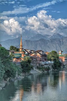 a river running through a lush green hillside next to a town with mountains in the background