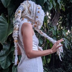 a woman wearing a white dress and crocheted headdress standing in front of trees