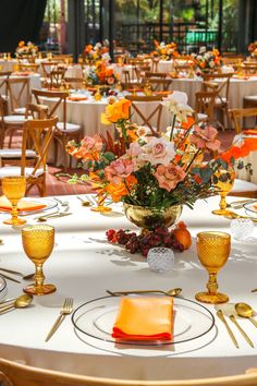 the table is set for an event with orange and white flowers in vases, silverware, and gold place settings