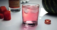 a pink drink with ice and watermelon next to it on a white table