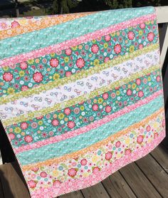 a multicolored quilt is hanging on a wooden porch railing with flowers and stripes