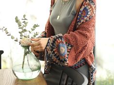 a woman sitting at a table holding a vase with flowers in it and a cardigan over her shoulder