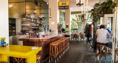 people are sitting at tables in a restaurant with yellow stools and green plants hanging from the ceiling