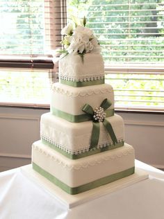 a three tiered wedding cake with green ribbon and flowers on top, sitting in front of a window