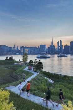 people are walking along a path near the water in front of a large city skyline