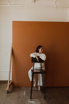 a woman sits on a chair in front of an orange wall and looks off to the side