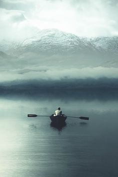 a man in a row boat on a lake with mountains in the background and foggy sky