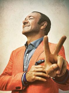 a man in an orange jacket making the v sign with his hand while wearing a blue shirt and tie