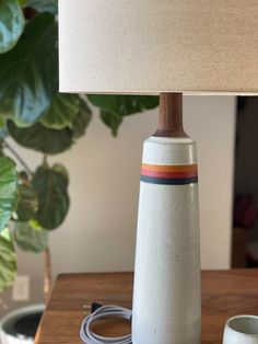 a table lamp sitting on top of a wooden table next to a white vase with a red, orange and blue stripe