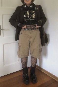 A mid-sized person/girl wearing a    black cropped graphic shirt, wide brown cargo shorts, black skull headphones, black cobweb tights, platform dr. Martens and multiple dark green and gray crystal rings and bracelets. 		The person is also wearing multiple silver necklaces. Masc Goth Outfits Plus Size, Fairycore Outfit Masc, Cottage Punk Aesthetic, Witch Core Outfits Male, Masc Goth Outfits Summer, Plus Size Alt Fashion Masc, Goblincore Masc Outfit, Crowcore Outfit Masc, Forest Grunge Outfit Masc