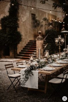 a long table with white linens and greenery is set for an outdoor dinner