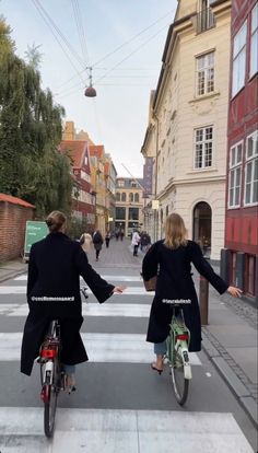 two people riding bikes down the street in front of some buildings and one person is pointing at something