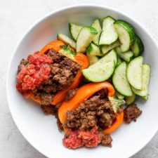 a white bowl filled with meat and veggies next to cucumbers on a table