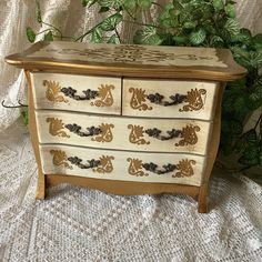 a white and gold painted chest of drawers on a lace covered tablecloth next to a potted plant