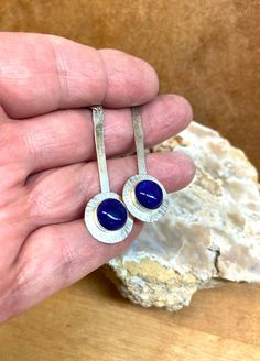 10mm Lapis Lazuli cabochons are bezel set on a larger Argentium Sterling silver disc, which is soldered to a hammered wire to make a beautiful dangly earring set.  Drilled holes at the tip of the hammered wires hold the ear wires.  The round silver disc is textured with hash marks around its perimeter.  They hang 2.5 inches with the ear wire being .75 inches of that. These beauties have definitely a feminine vibe with a modern feel. Argentium sterling silver is a recycled metal with more fine silver than regular sterling silver, but an added bit of germanium to help make it tarnish resistant.  All my metal and wire is Argentium 35. Lapis is noted for being a powerful stone for thinking and spirituality.  It is said to increase self-knowledge and awareness of your thoughts. Modern Sterling Silver Earrings With Cabochon, Hand Forged Blue Round Earrings, Round Metal Cabochon Earrings, Blue Hammered Earrings For Gift, Sterling Silver Bezel Set Earrings, Sterling Silver Bezel Set Round Earrings, Round Bezel Set Cabochons For Gifts, Dangly Earring, Industrial Earrings