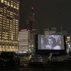 an outdoor movie screen in the middle of a parking lot with cars parked around it